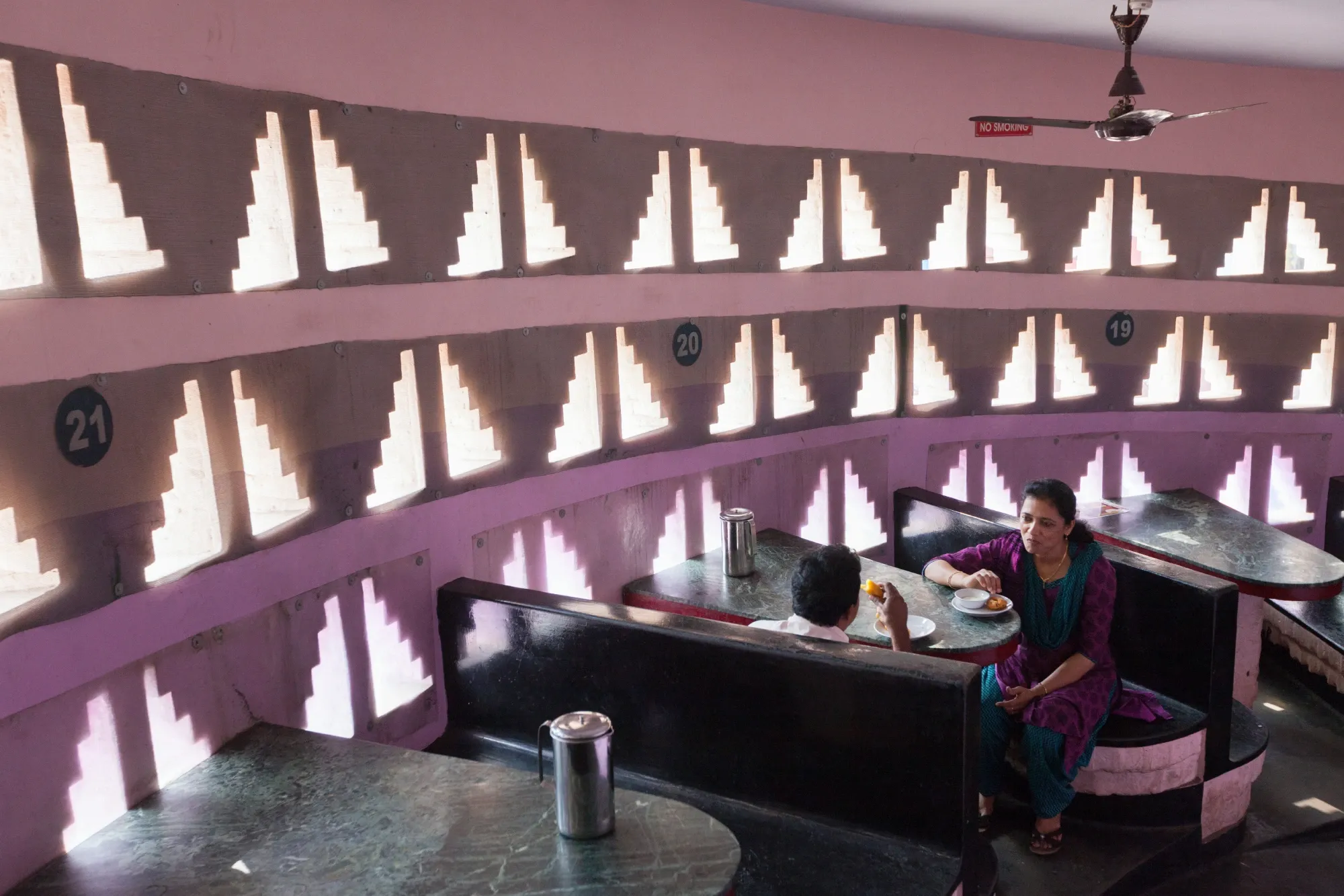 Two people sit in a booth talking, the wall beside them is full of geometric-shaped openings