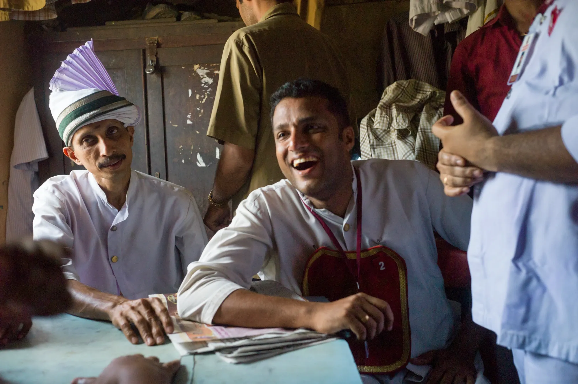 Two people dressed in white waiters' uniforms sit at a table chatting and laughing with someone behind the camera