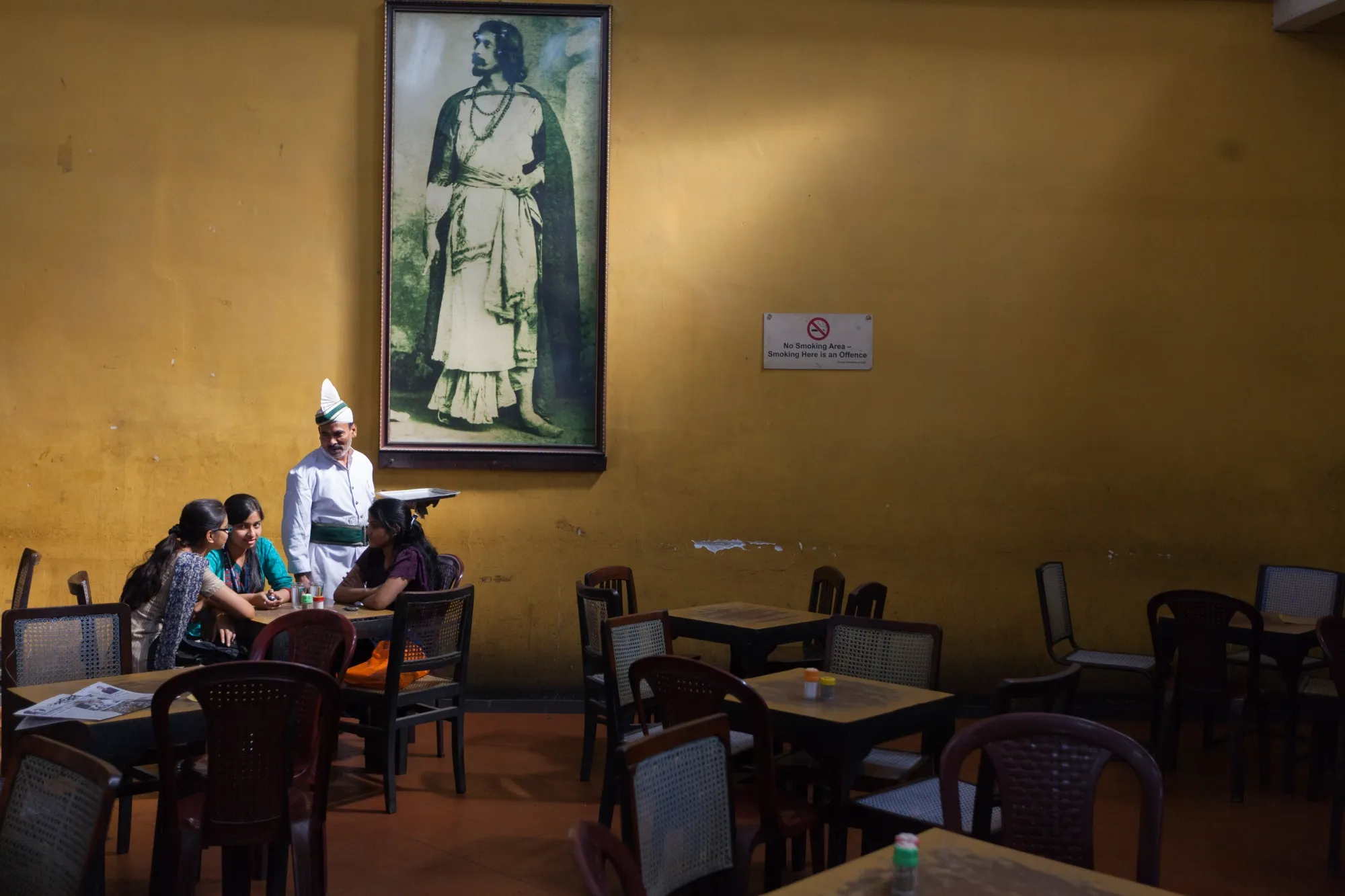 Wide shot of a cafe interior, three people sit at a table being served by a waiter. A large portrait hangs on the wall behind them.