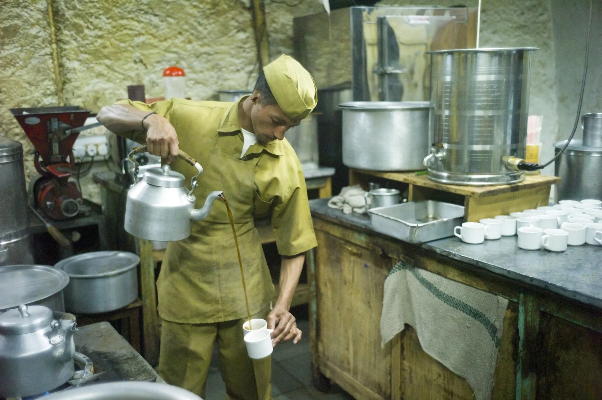 A worker expertly pours coffee from a big kettle from a height into two cups in a coffee house kitchen