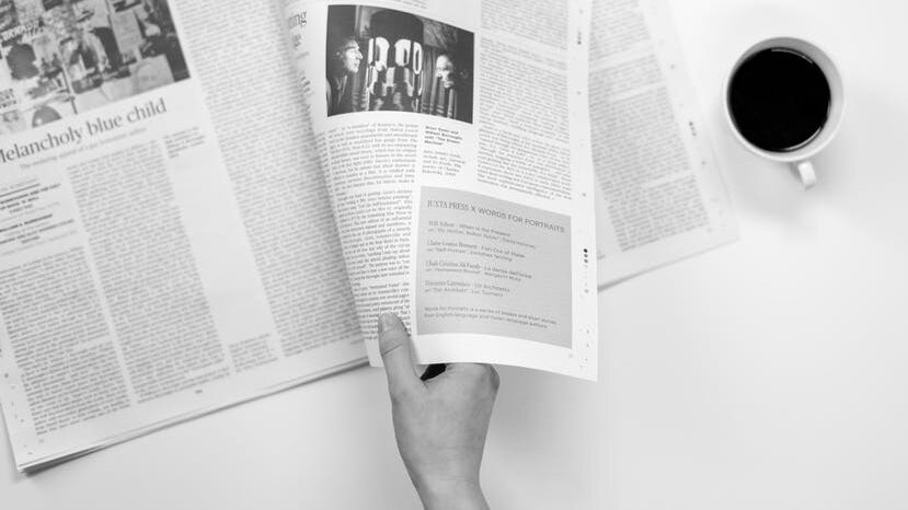 A hand turns the pages of a newspaper next to a cup of coffee, seen from above. Via Pexels.