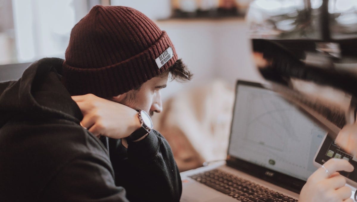Over the shoulder of a person in a beanie hat resting on their arm while looking at a computer screen