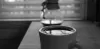 A cup of coffee on a table in the foreground with a Chemex out of focus in the background.